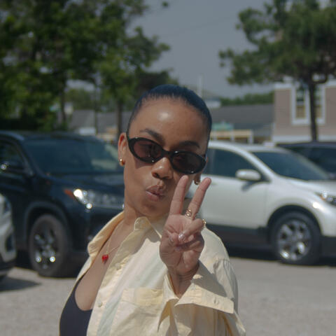 Dre Pedemonte A young woman wearing sunglasses and a light yellow shirt poses playfully outdoors, holding up a peace sign. Cars and trees are visible in the sunny background.